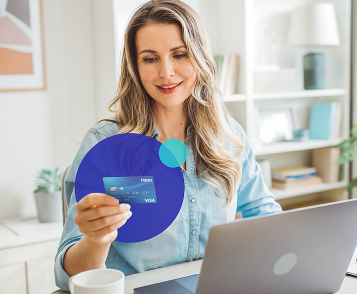 donna bionda sorridente seduta a una scrivania con laptop, tiene in mano una carta di credito blu Nexi. L'ambiente è domestico e luminoso con scaffali bianchi sullo sfondo. Un elemento grafico blu e turchese sovrapposto suggerisce l'e-commerce.