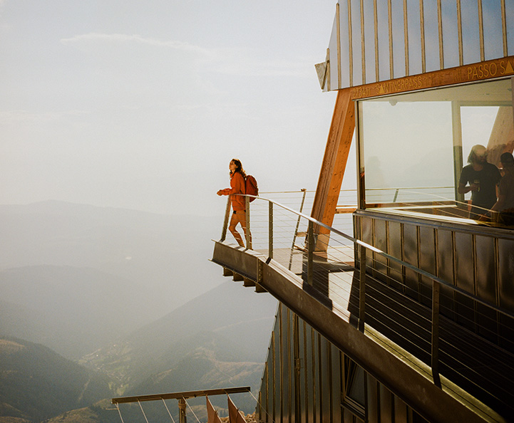 donna bionda sorridente seduta a una scrivania con laptop, tiene in mano una carta di credito blu Nexi. L'ambiente è domestico e luminoso con scaffali bianchi sullo sfondo. Un elemento grafico blu e turchese sovrapposto suggerisce l'e-commerce.